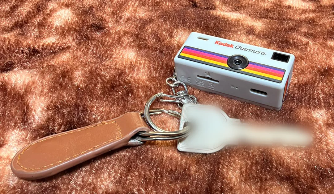 A small grey Kodak Charmera with a rainbow stripe, clipped to a keyring beside a house key and a leather fob.