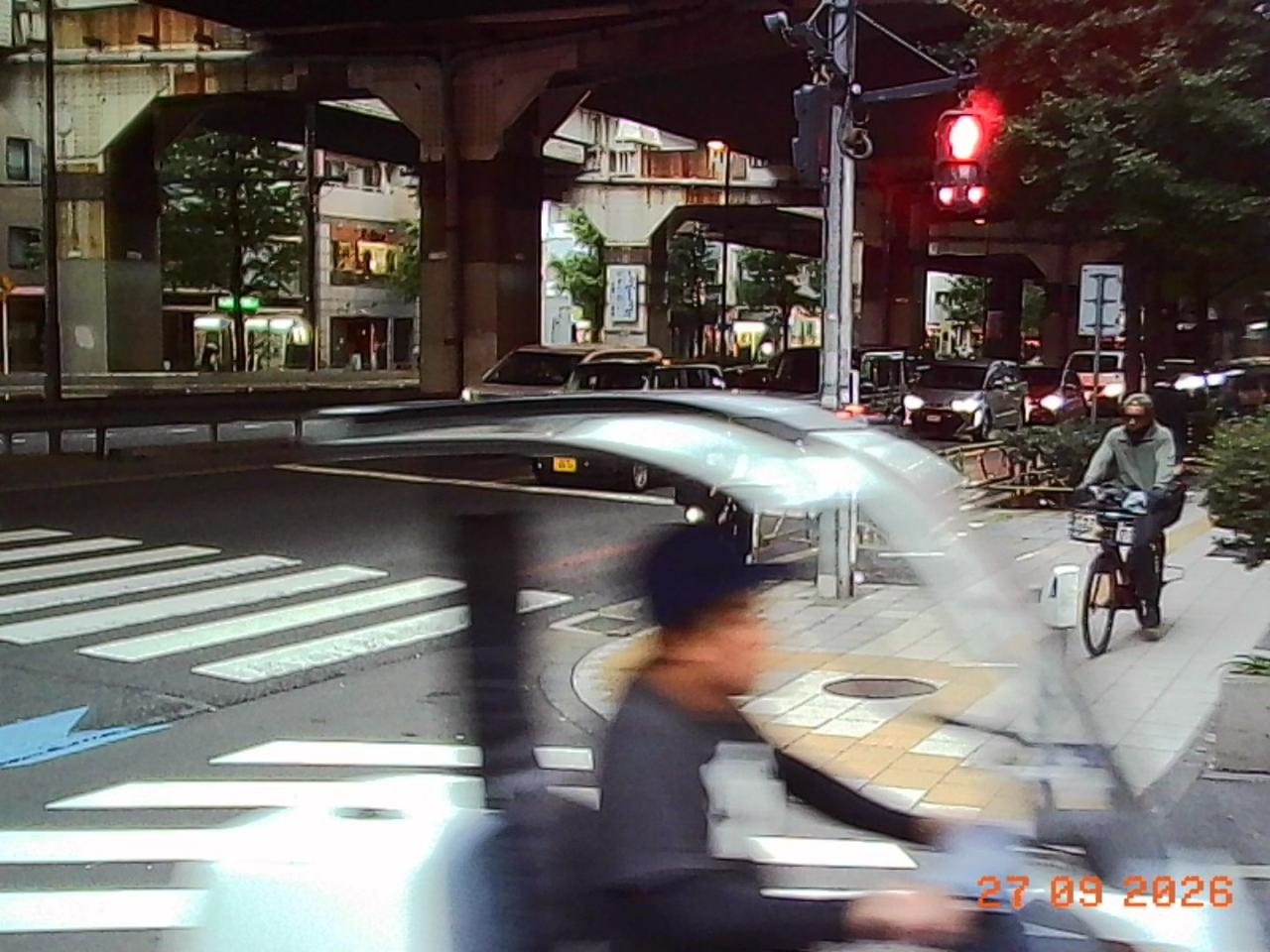 Charmera photo: under an elevated road in Tokyo, a pedestrian light glows red while a cyclist in the foreground smears past; the date stamp reads 27 09 2026.